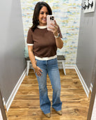 A woman takes a mirror selfie in a fitting room wearing a brown short-sleeve knit top with cream ribbed trim and light blue flare jeans.
