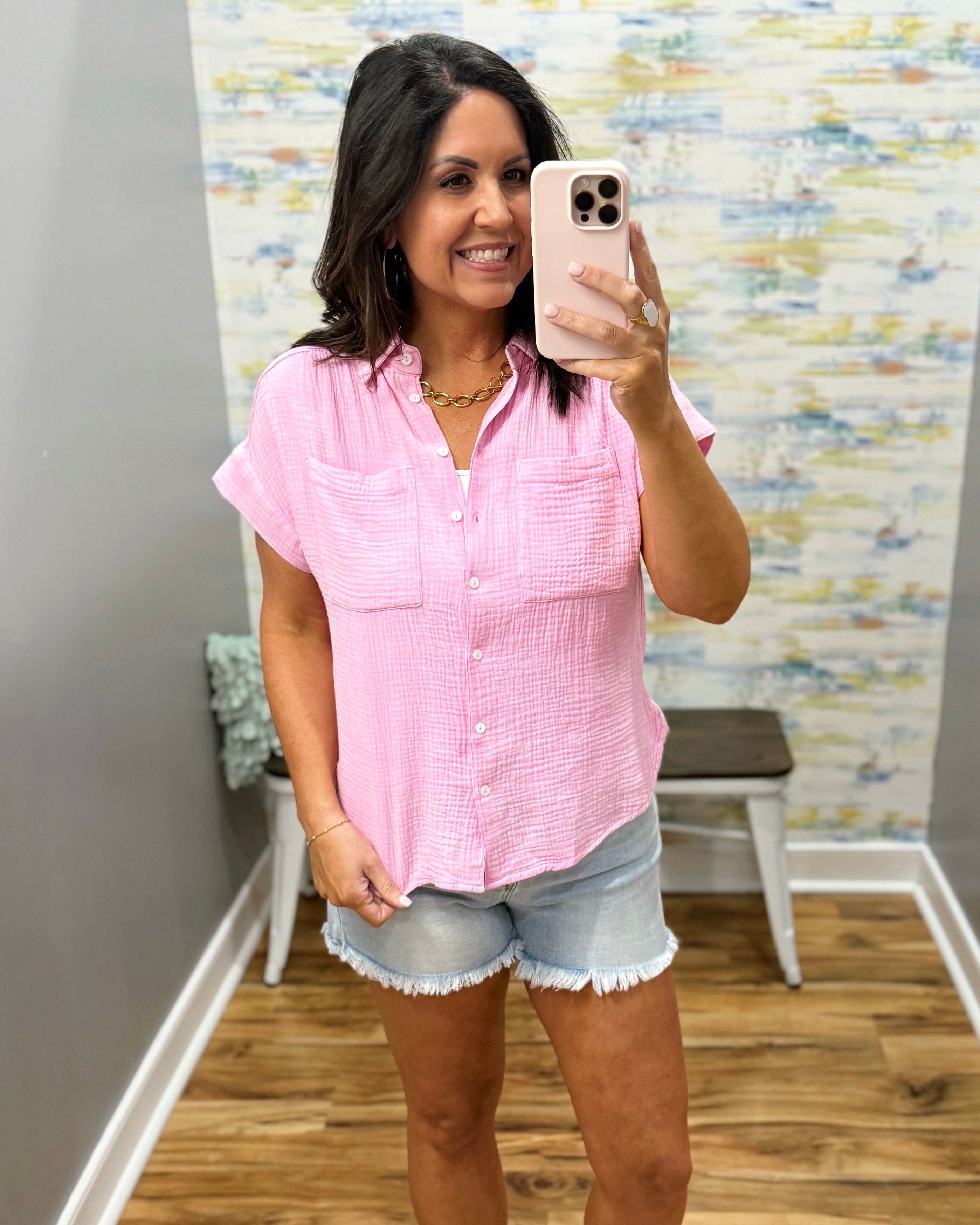 Woman taking a mirror selfie wearing a pink shirt and denim shorts in a room with a textured wall.