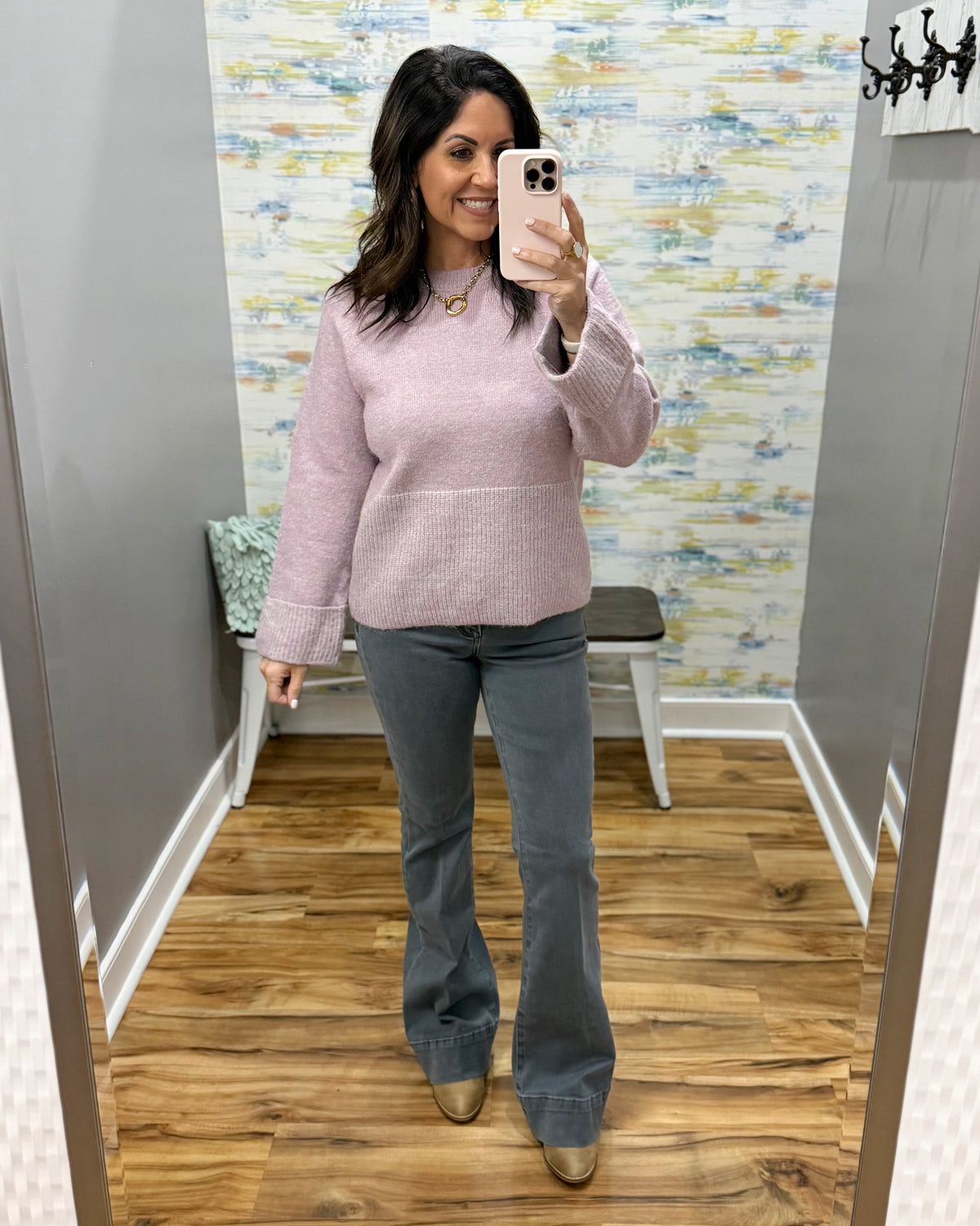 A woman takes a mirror selfie in a fitting room wearing a light pink knit mock-neck sweater and gray flared jeans with metallic shoes.