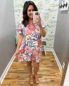 Woman taking a mirror selfie wearing a floral dress in a room with colorful wallpaper.