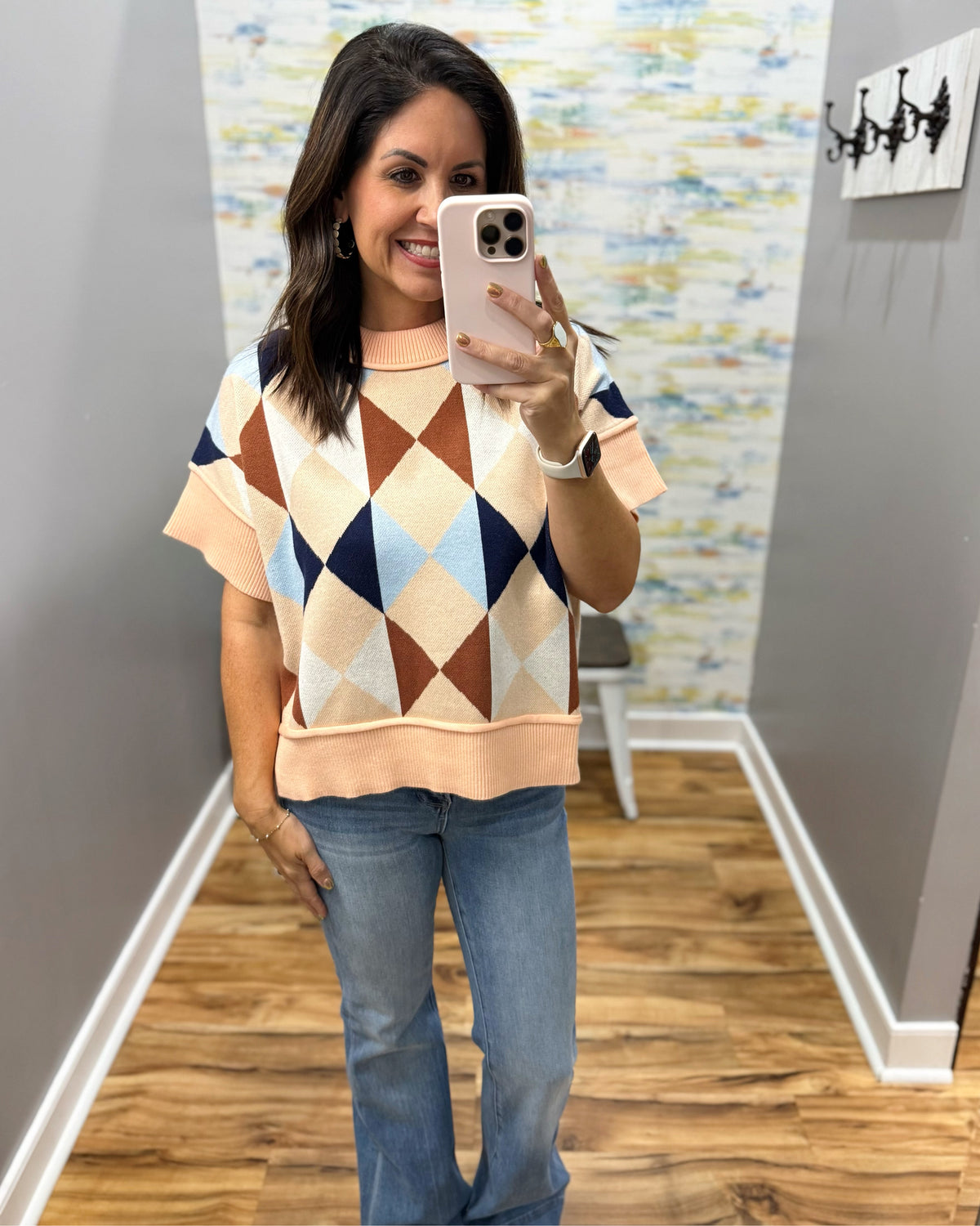 A woman takes a mirror selfie wearing a short-sleeve knit sweater with a multicolor argyle pattern in peach, cream, brown, and light blue, paired with blue jeans.