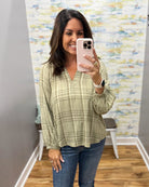 A woman takes a mirror selfie wearing a sage green and beige plaid long-sleeve blouse with blue jeans, standing in a room with a patterned wallpaper background.