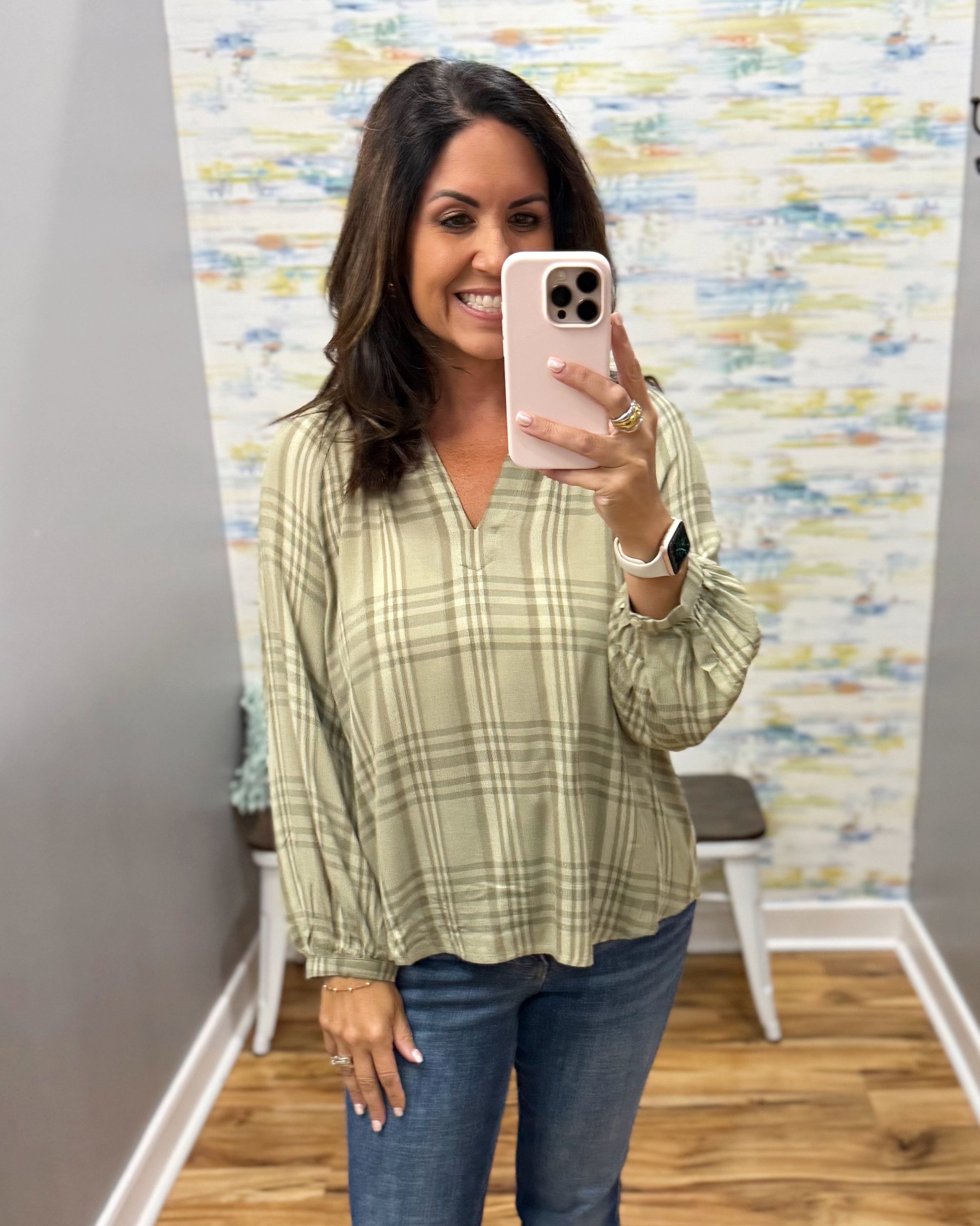 A woman takes a mirror selfie wearing a sage green and beige plaid long-sleeve blouse with blue jeans, standing in a room with a patterned wallpaper background.