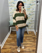 A woman takes a mirror selfie in a fitting room wearing a knit sweater with wide horizontal stripes in dark green and cream, paired with blue jeans and beige sneakers.
