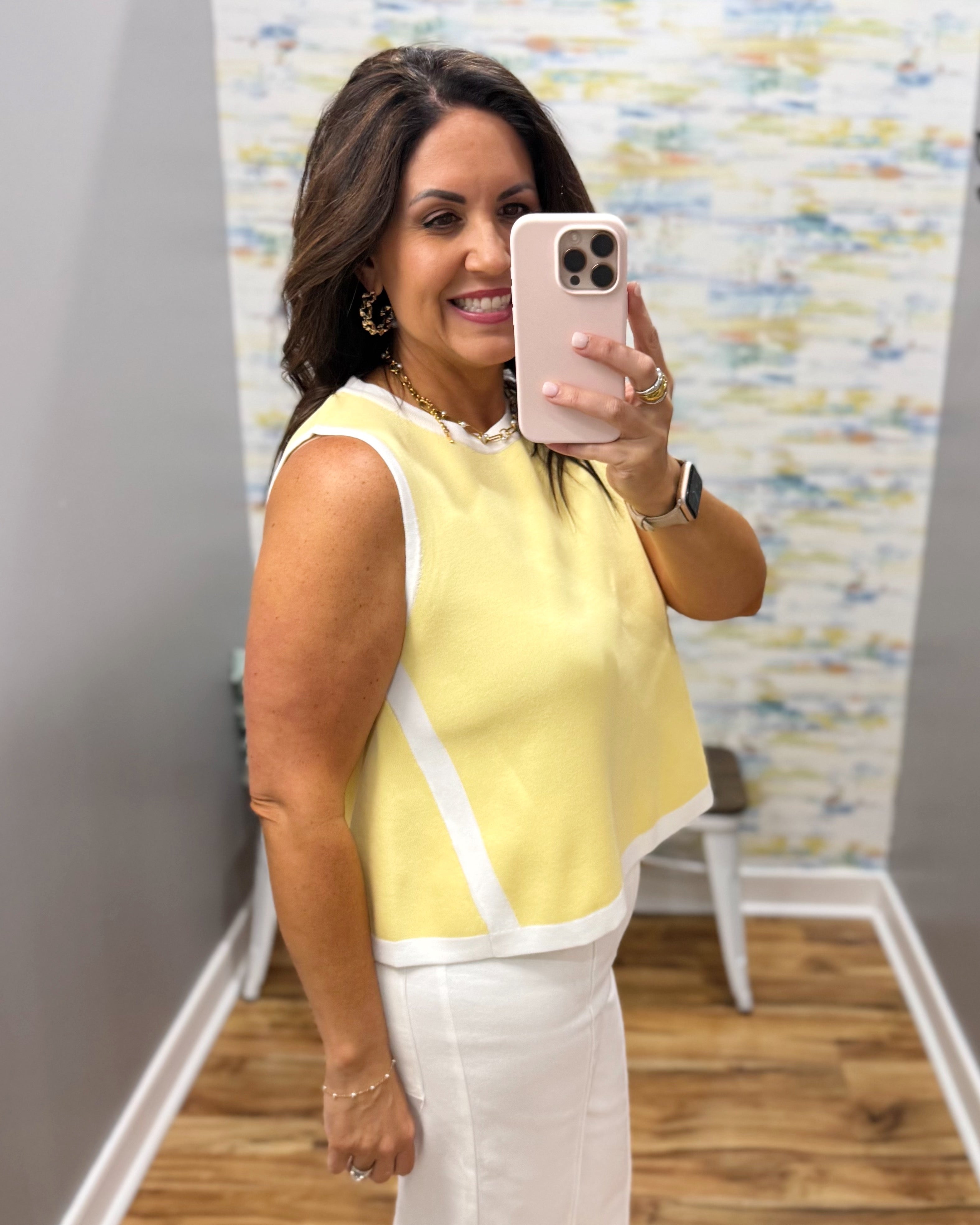 Woman taking a mirror selfie wearing a yellow top and white pants in a room with a colorful wall.