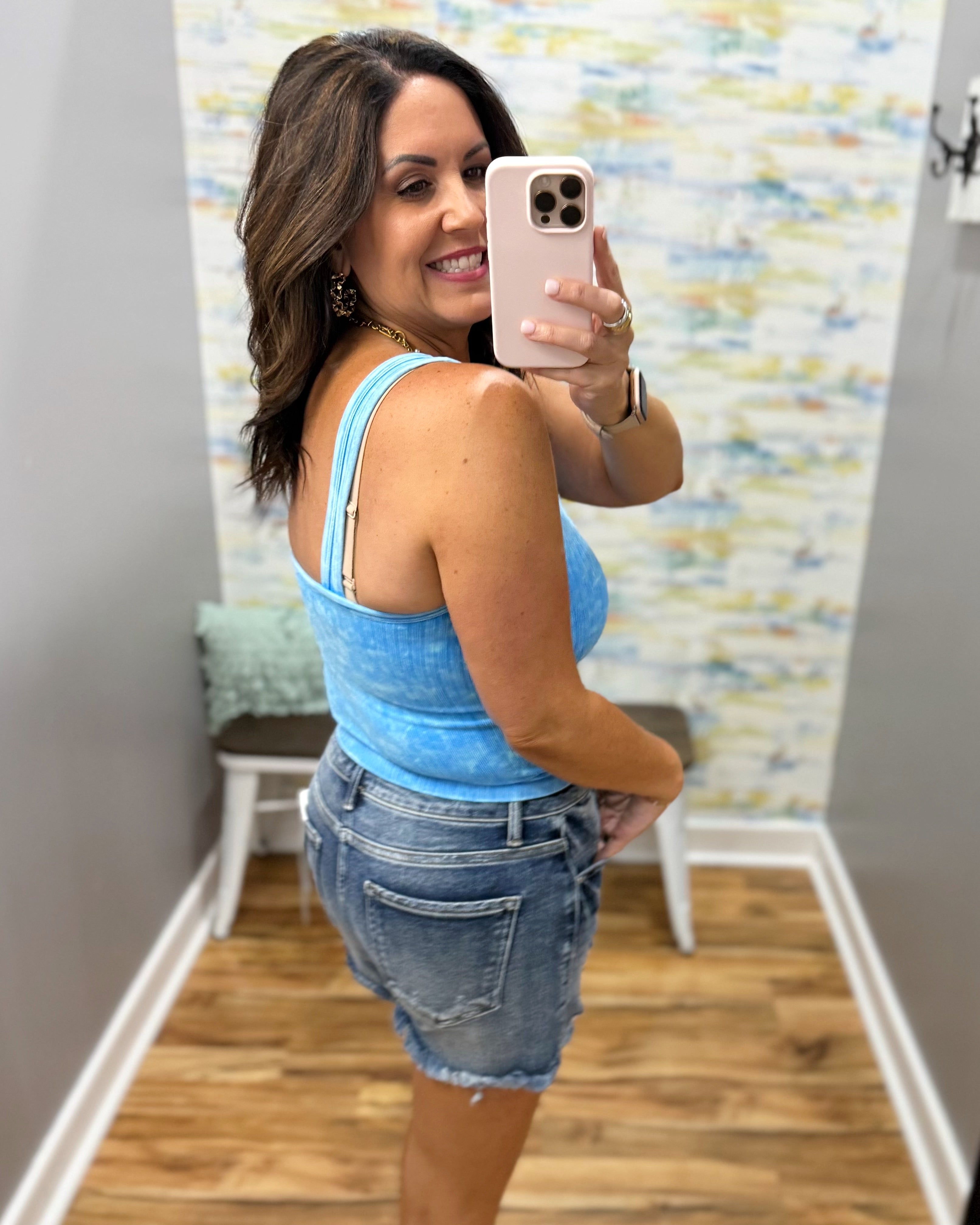 Woman taking a mirror selfie wearing a blue tank top and denim shorts in a room with a colorful wall.