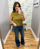 A woman takes a mirror selfie in a fitting room wearing a sleeveless olive green mock-neck knit top and blue flare jeans.