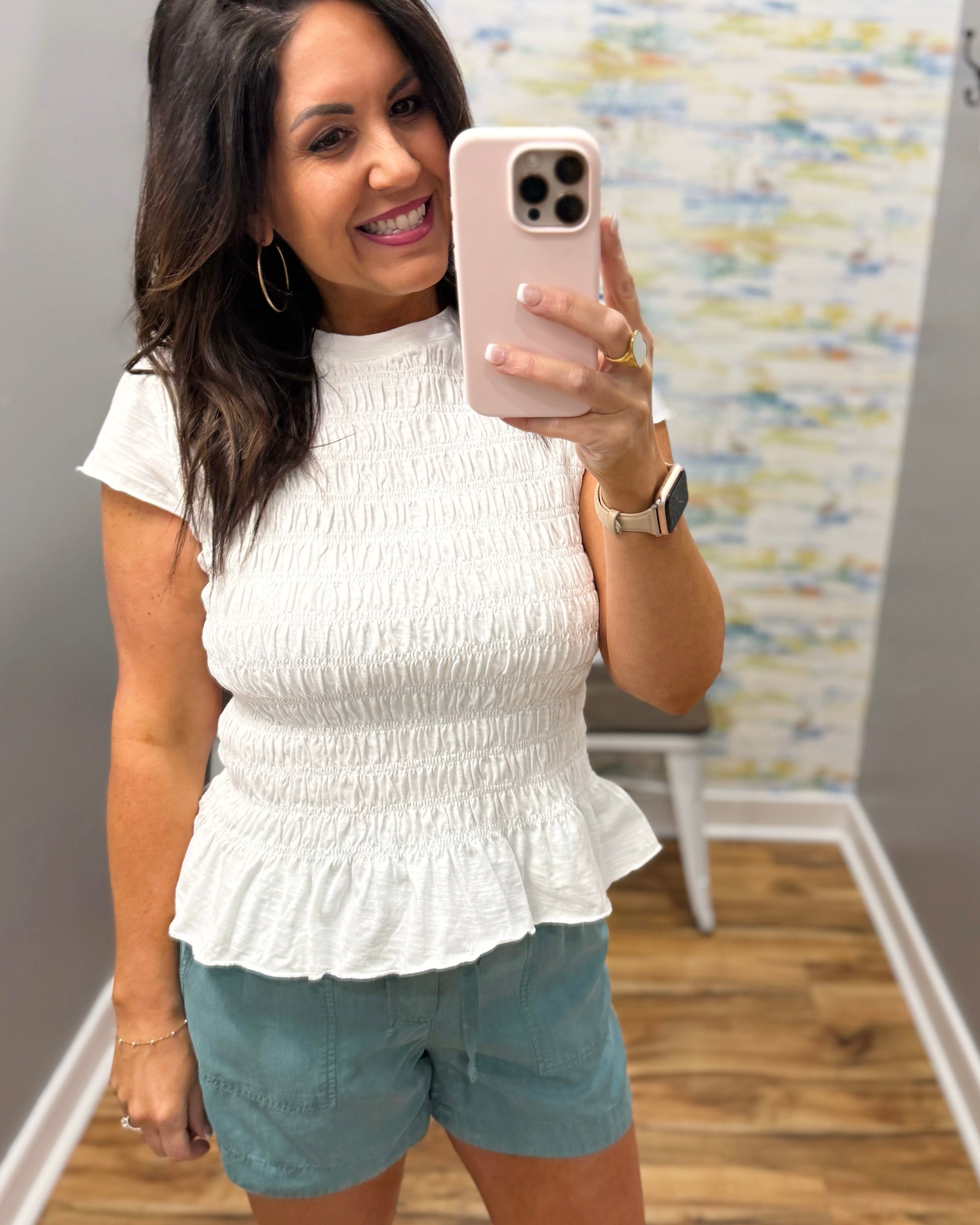 Woman taking a mirror selfie wearing a white ruffled top and light blue shorts.