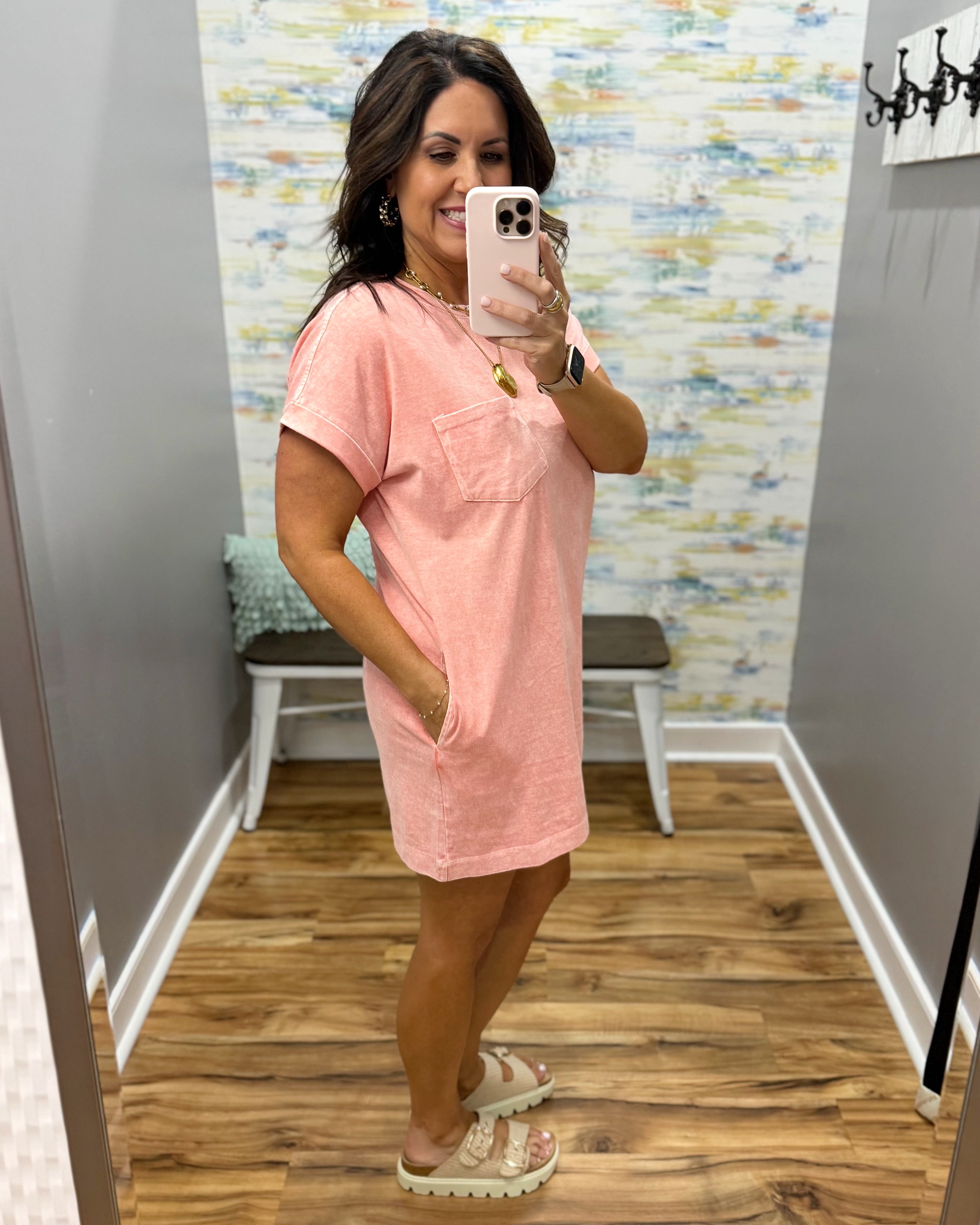 Woman taking a mirror selfie wearing a pink dress in a room with patterned wallpaper.