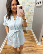 Woman wearing a light blue dress in a room with wooden flooring and a patterned wall.