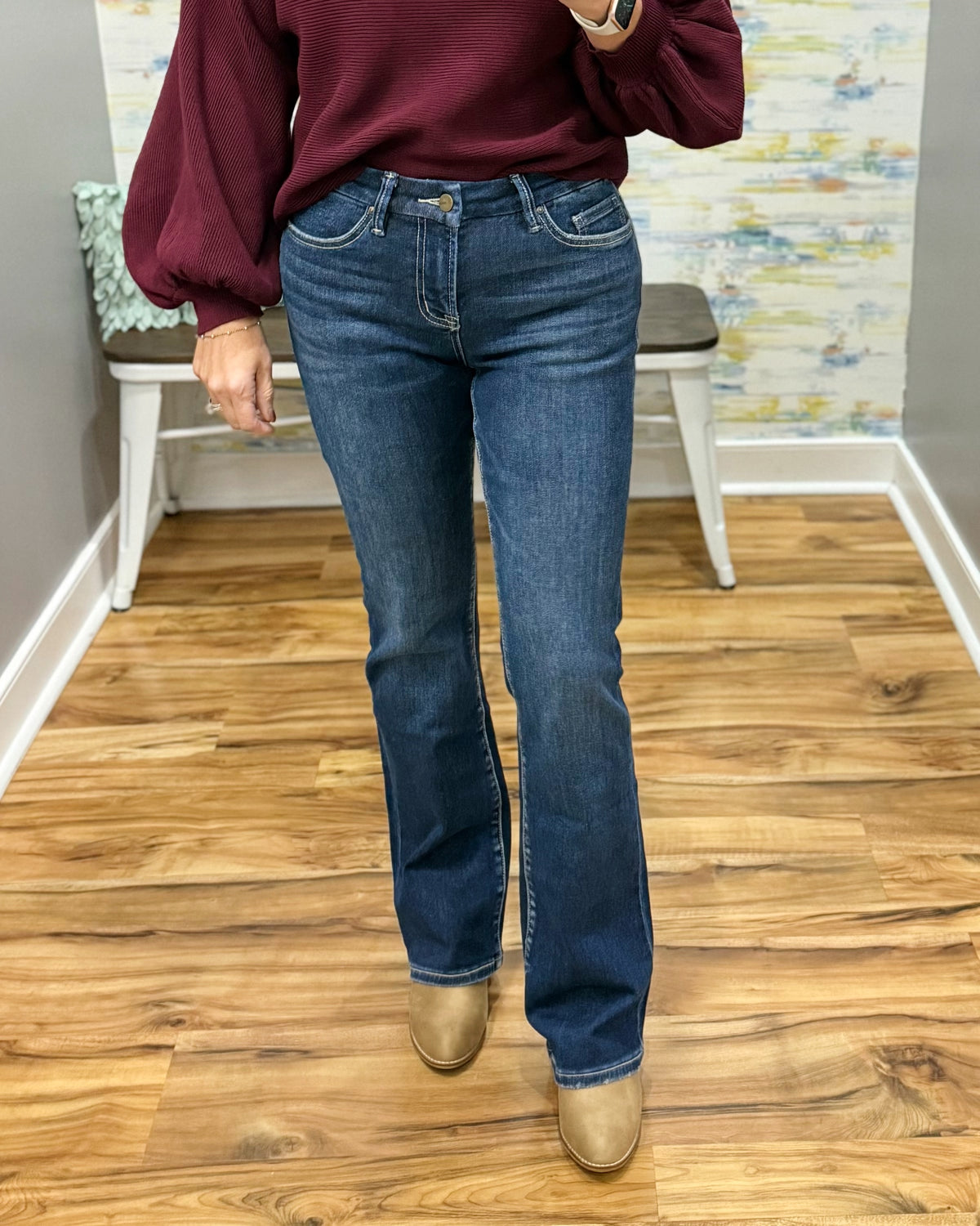 A person wearing mica dark blue bootcut jeans, a burgundy long-sleeve top, and tan boots, standing on a wood floor in front of a wall with a bench.