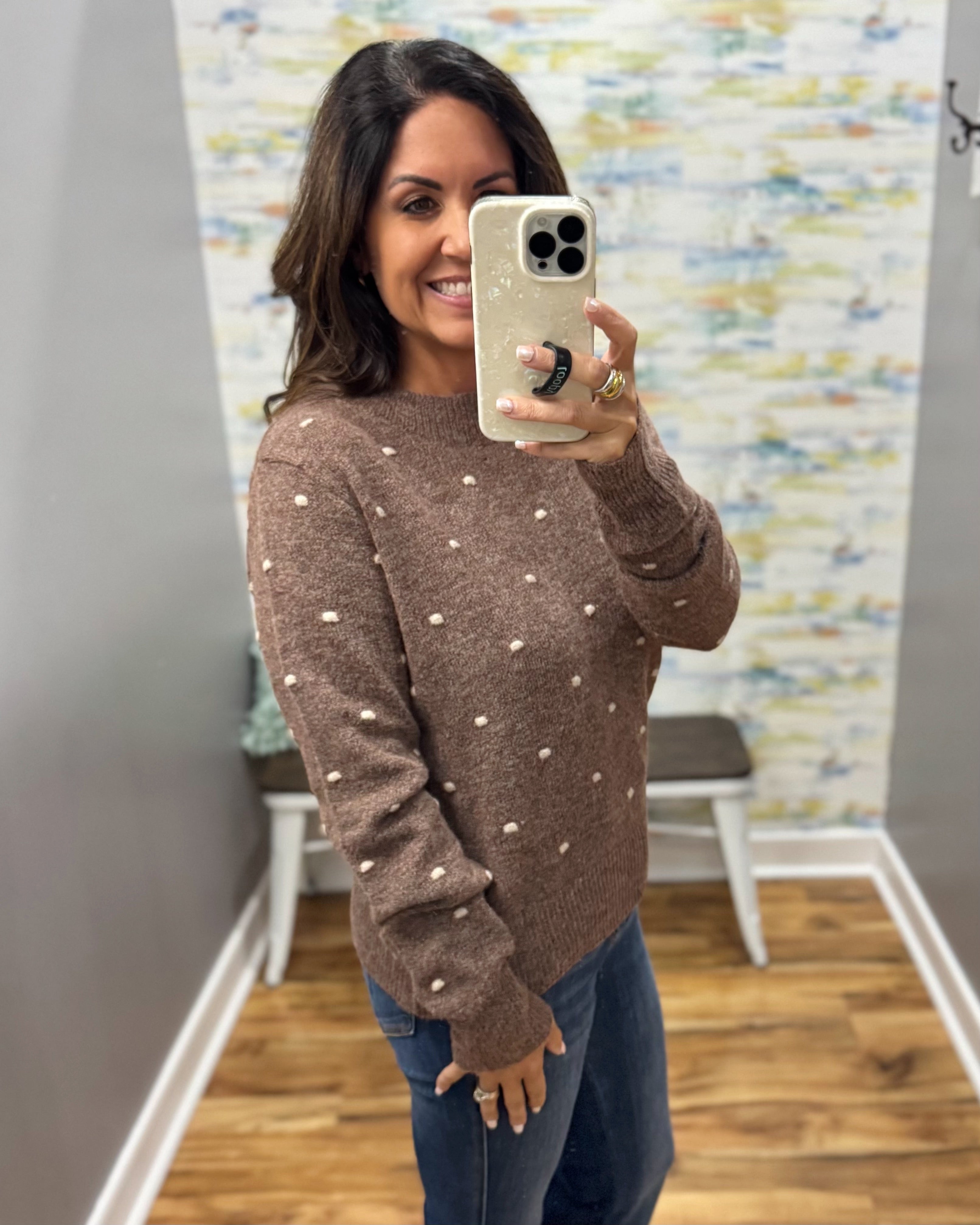 A woman takes a mirror selfie wearing a brown knit sweater with small white polka dots and blue jeans in a fitting room with wood floors and a patterned wall.