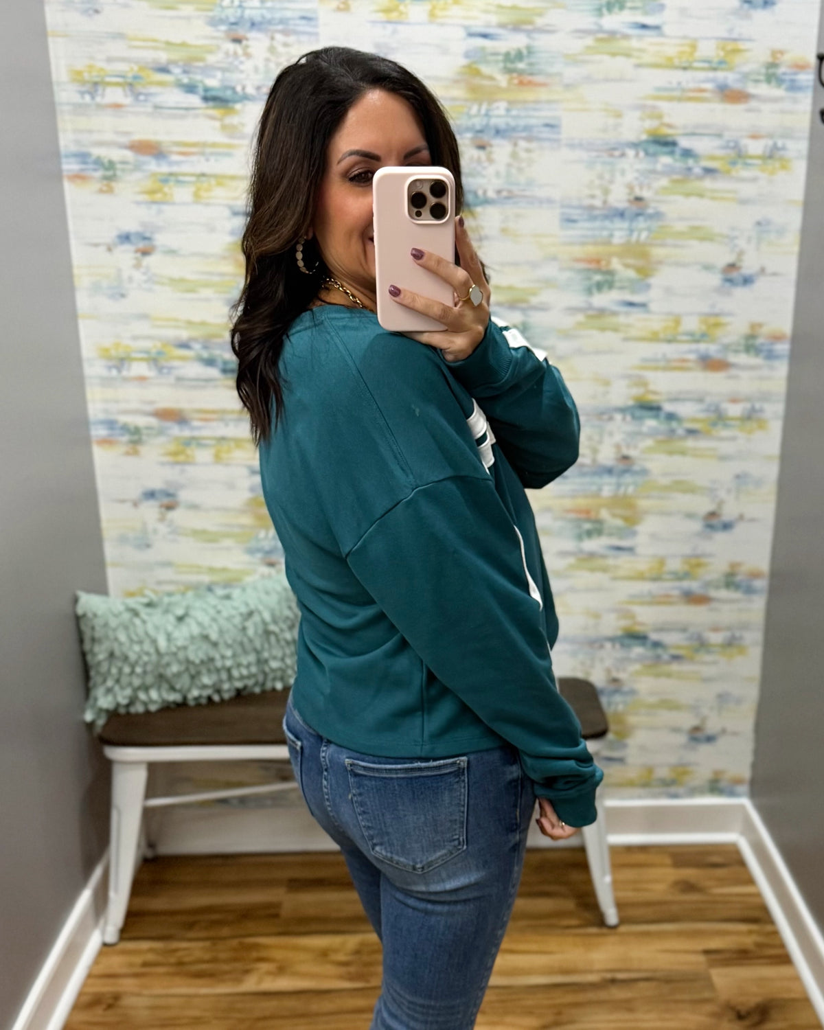 A woman taking a mirror selfie in a fitting room wearing a teal green sweatshirt and blue jeans, with a patterned wallpaper background and a small bench with a pillow.