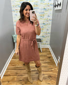 A woman takes a mirror selfie wearing a short-sleeve dusty rose knit dress with a tie waist and tan knee-high boots in a home setting with wood floors and patterned wallpaper.