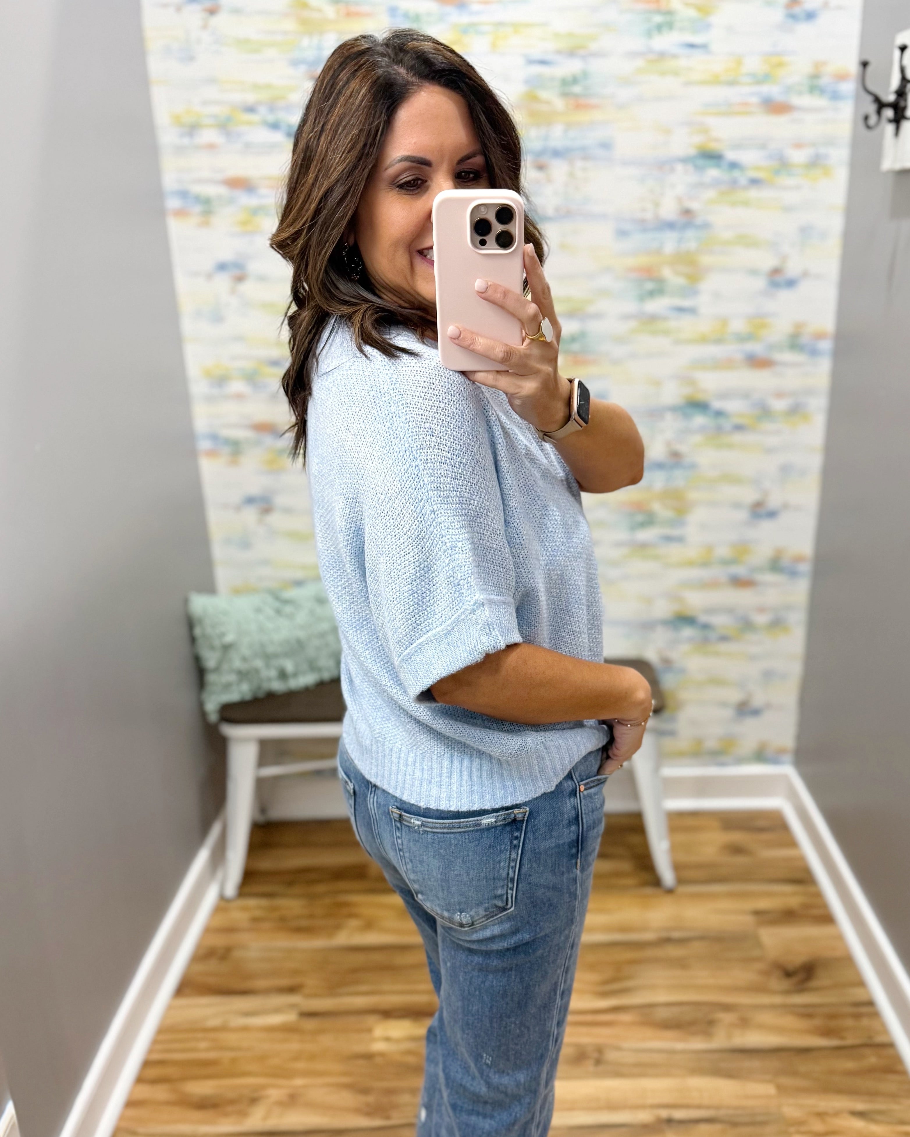 Woman taking a mirror selfie wearing a light blue shirt and jeans in a room with a colorful wall.