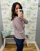 A woman taking a mirror selfie wearing a long-sleeve crewneck top with horizontal burgundy and white stripes, paired with dark blue jeans, in front of a patterned wallpaper background.