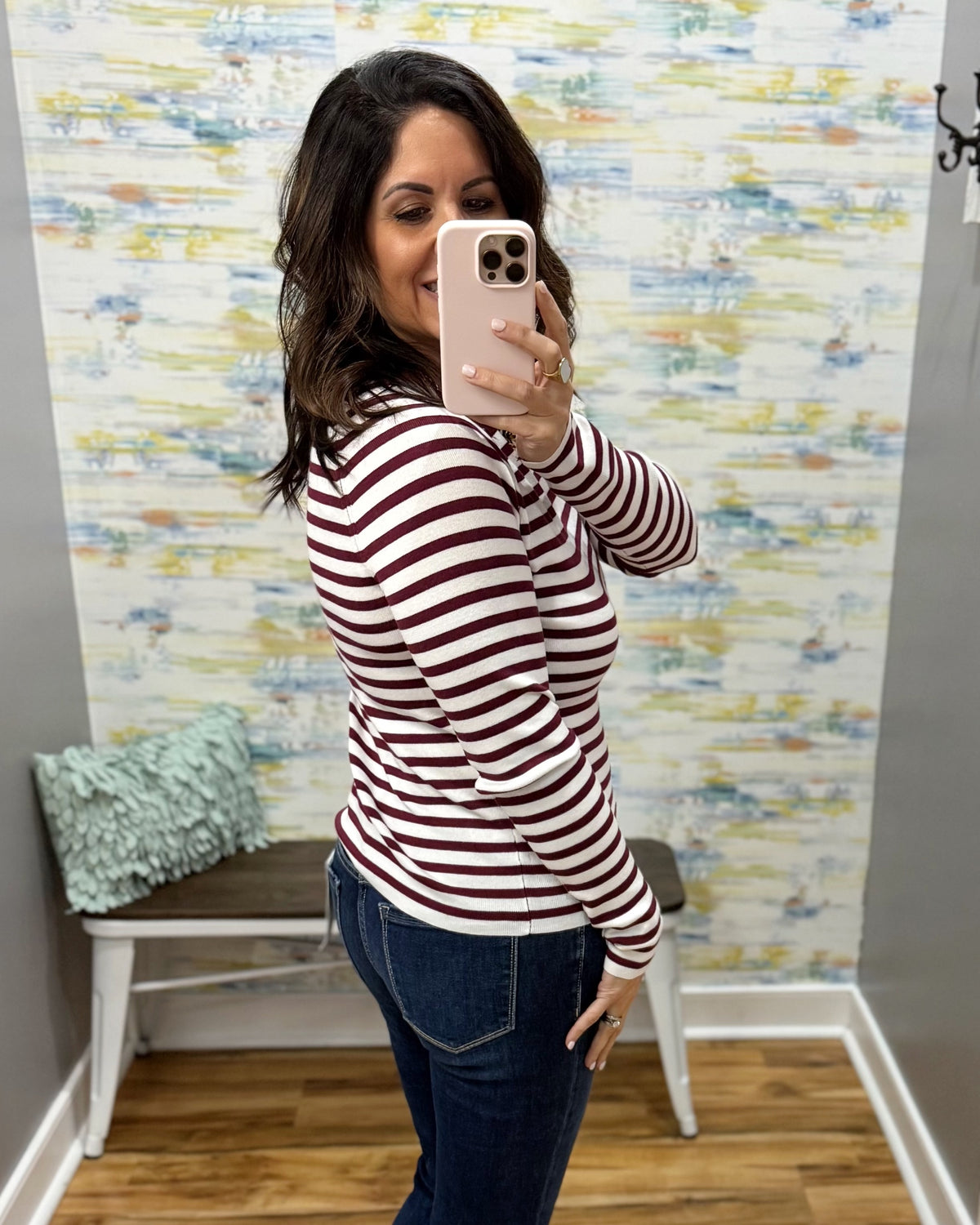 A woman taking a mirror selfie wearing a long-sleeve crewneck top with horizontal burgundy and white stripes, paired with dark blue jeans, in front of a patterned wallpaper background.