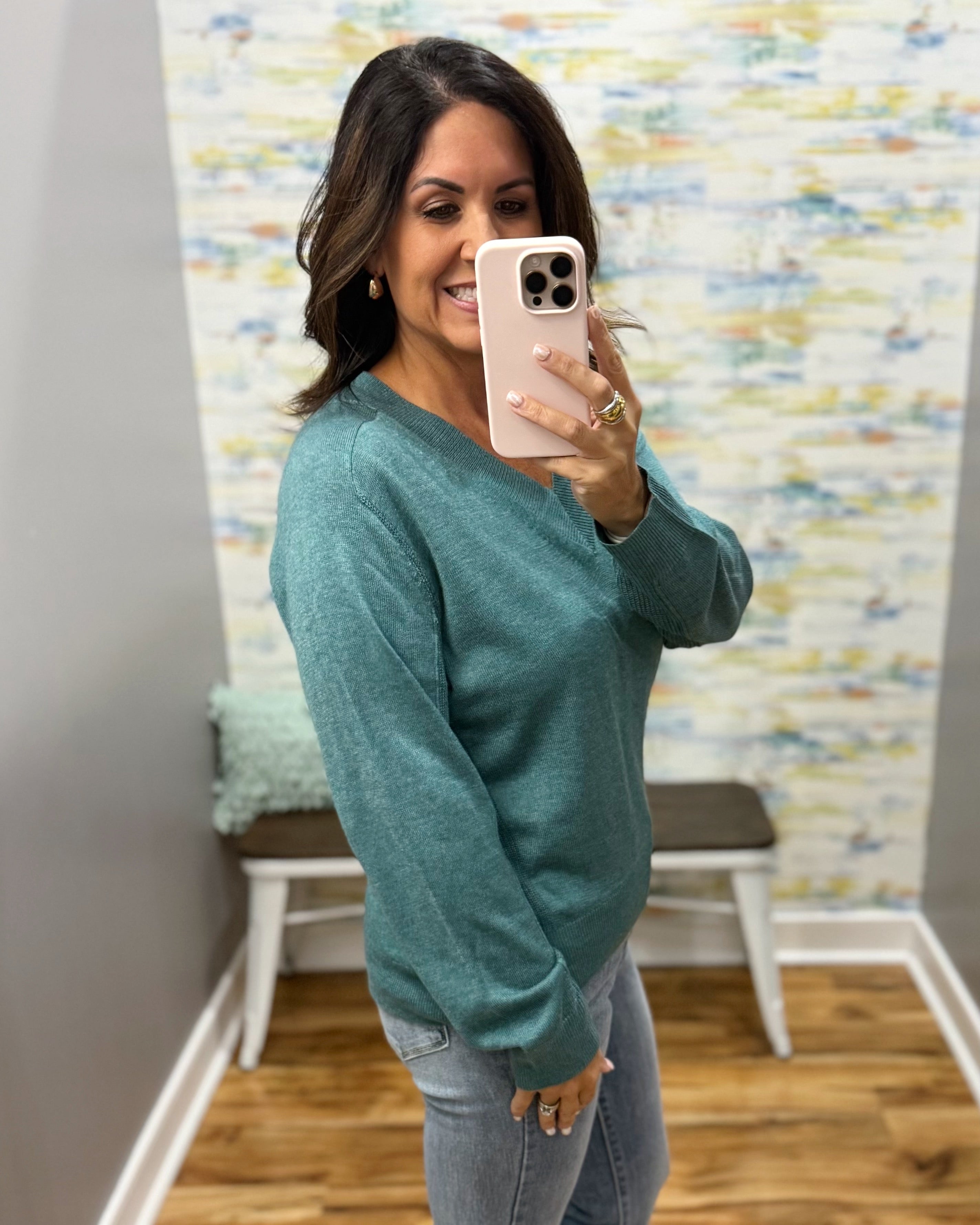 A woman takes a mirror selfie wearing a heathered teal green long-sleeve sweater and light wash jeans in a home setting with a patterned wallpaper background.