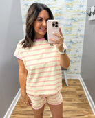 A woman takes a mirror selfie wearing a matching two-piece pajama set with light pink and white horizontal stripes. The set includes a short-sleeve crewneck t-shirt and matching shorts, shown in a home or boutique fitting room environment.