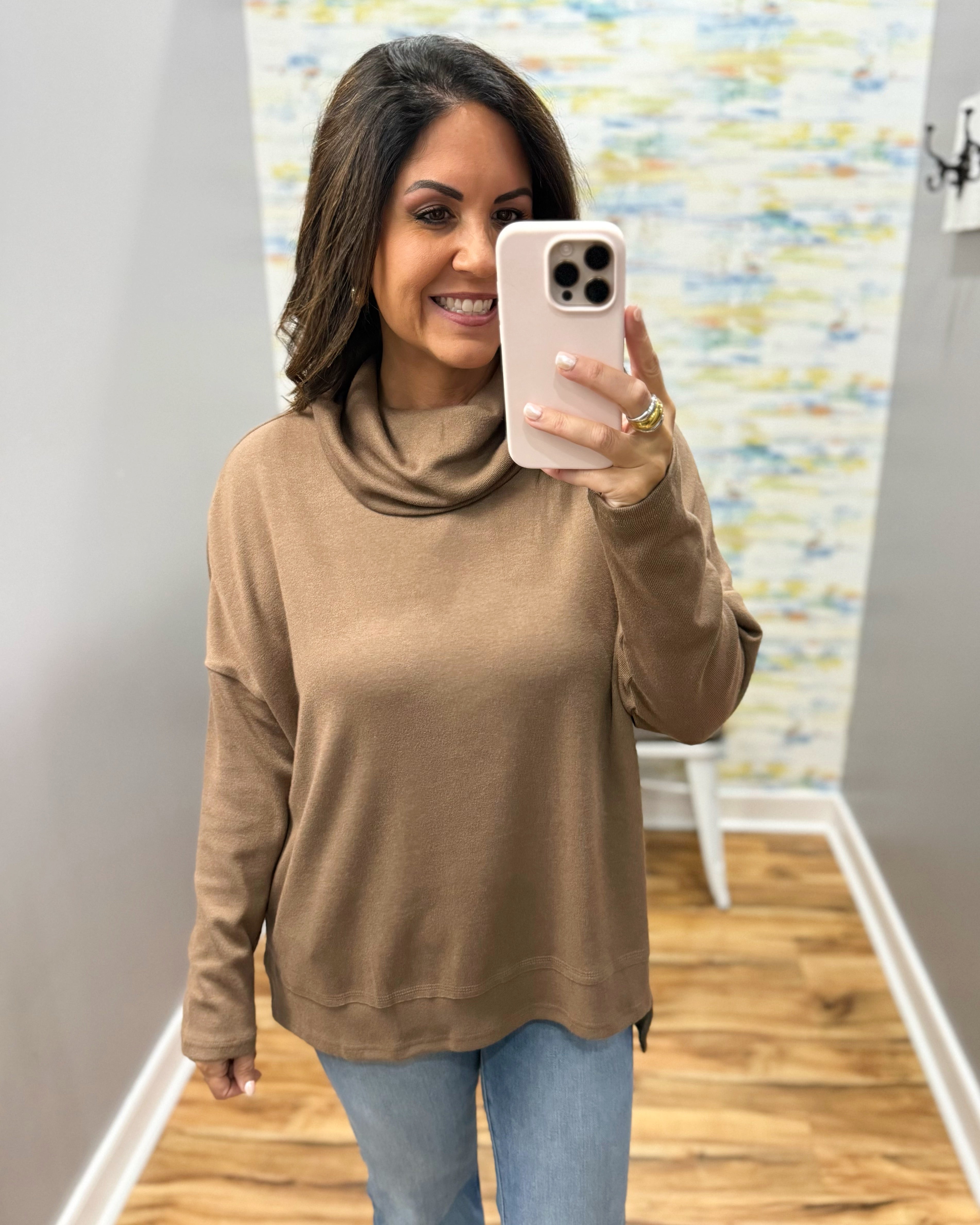 A woman takes a mirror selfie wearing a tan cowl-neck long-sleeve knit top and light blue jeans in an indoor hallway.