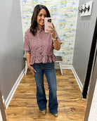 A woman takes a mirror selfie in a fitting room wearing a red and white gingham short-sleeve button-up shirt with a ruffled hem and blue flare jeans.