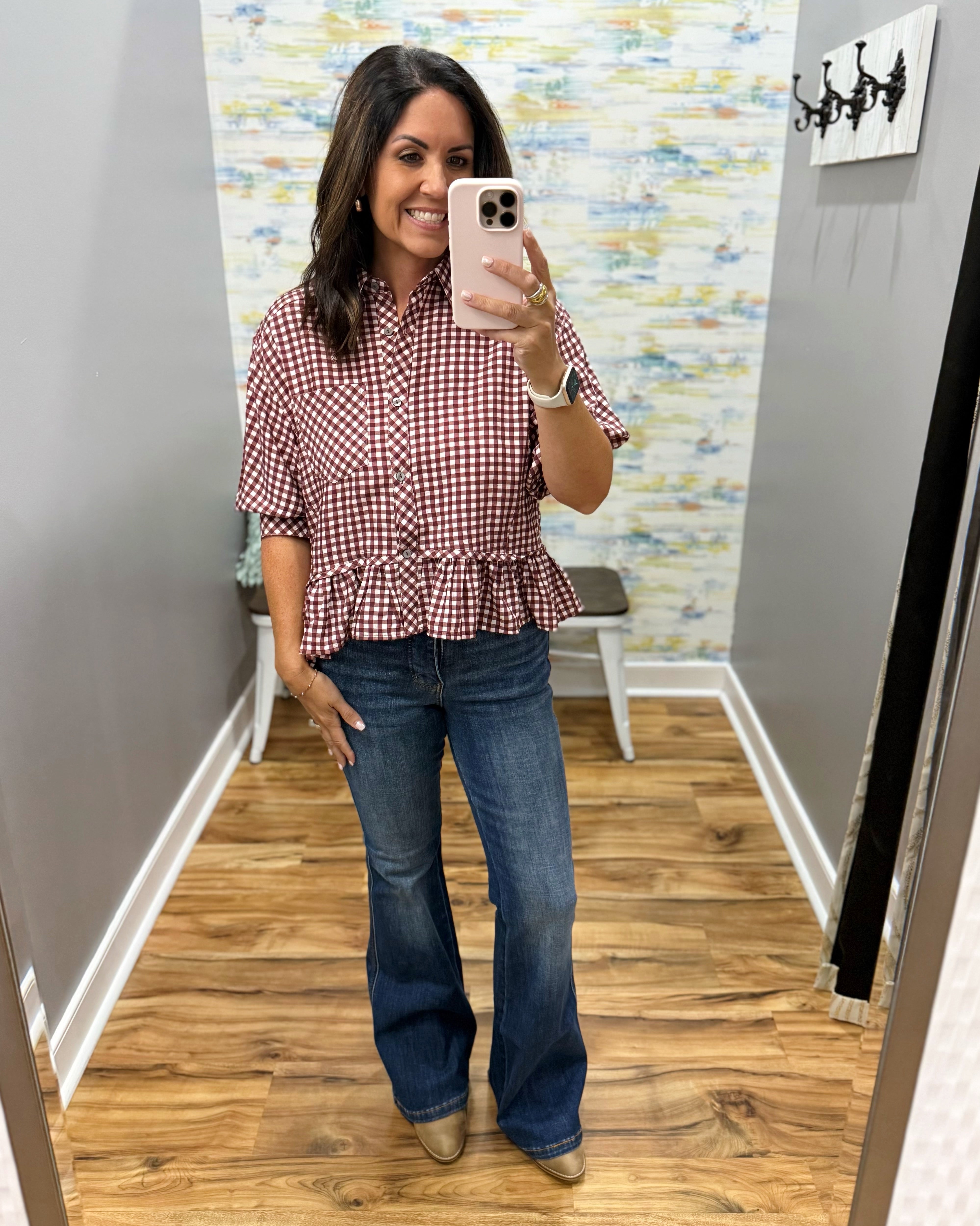 A woman takes a mirror selfie in a fitting room wearing a red and white gingham short-sleeve button-up shirt with a ruffled hem and blue flare jeans.