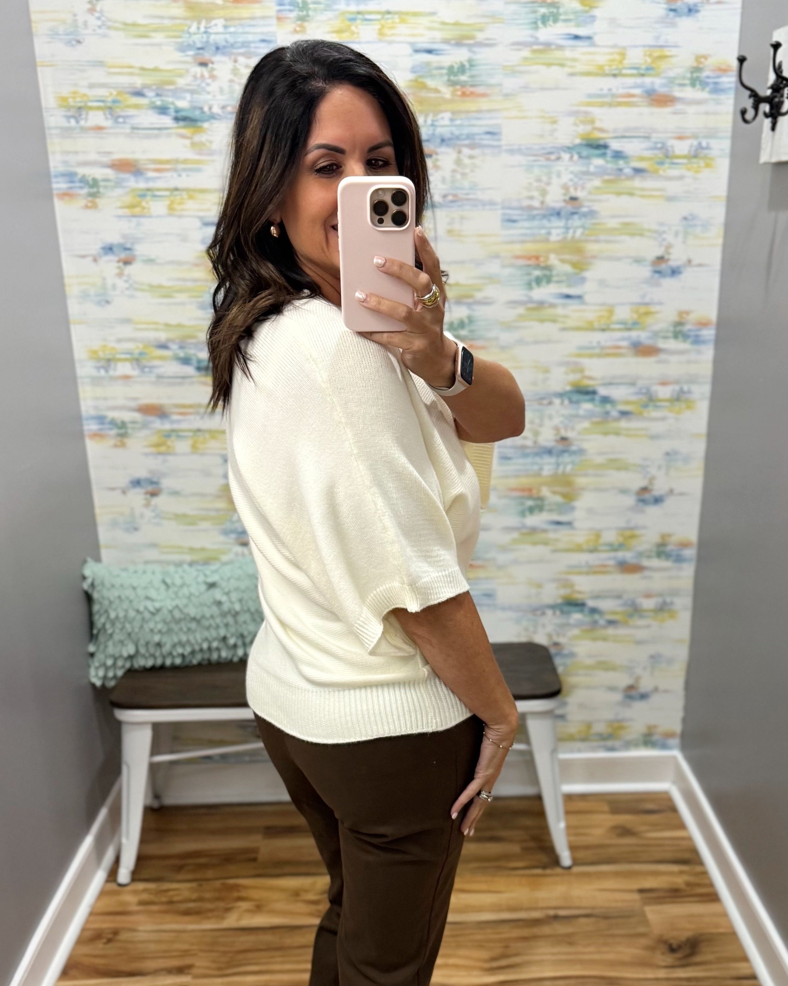 A woman takes a mirror selfie in a fitting room wearing a cream-colored short-sleeve knit top and dark brown pants. The background features a colorful abstract patterned wallpaper, a wooden bench with a green pillow, and light wood flooring.