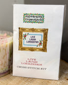 A packaged cross stitch kit labeled 'subversive cross stitch' and 'LIVE LAUGH LORAZEPAM' in a white box with a gold frame illustration and floral accents, photographed on a tabletop next to a candle.