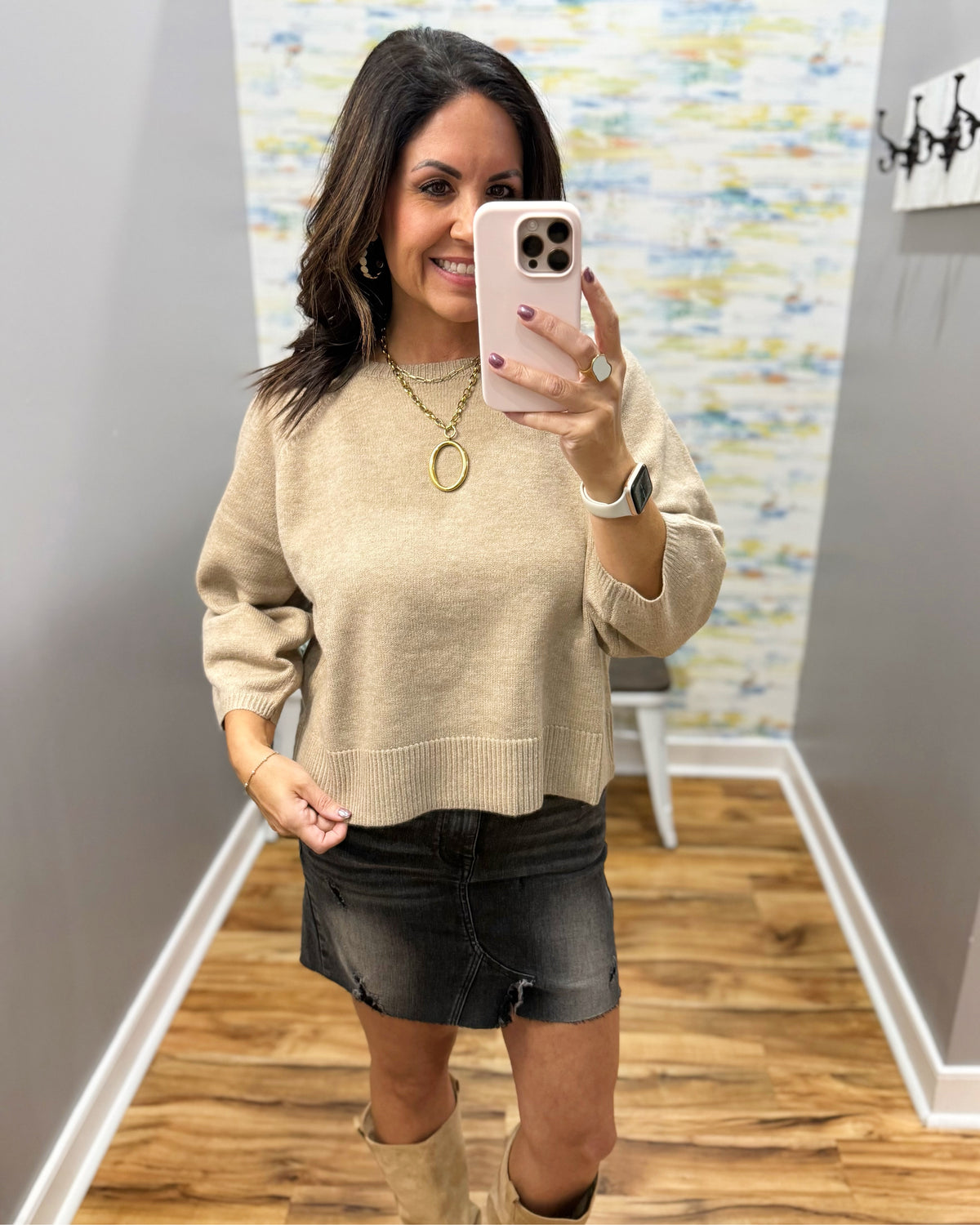 A woman taking a mirror selfie in a fitting room wearing a beige knit crewneck sweater, a black denim mini skirt, and beige ankle boots.
