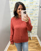 A woman takes a mirror selfie wearing a heathered rust-red knit sweater and light-wash blue jeans in a home setting with wood floors and a patterned wallpaper background.