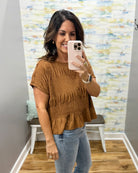 A woman takes a mirror selfie wearing a brown smocked short-sleeve peplum top and light blue jeans in a fitting room with a patterned wall backdrop.