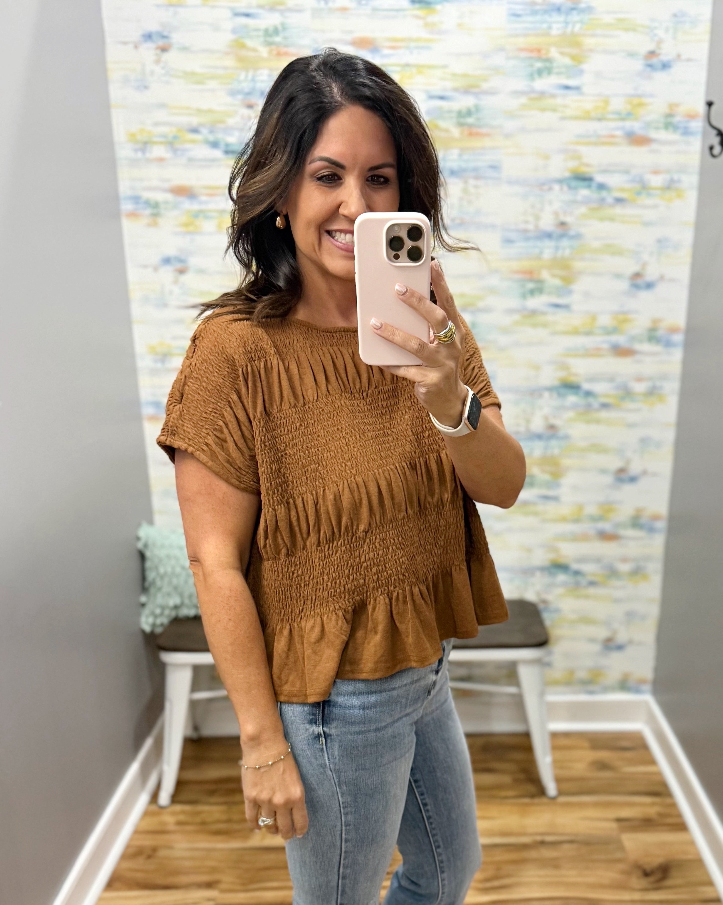 A woman takes a mirror selfie wearing a brown smocked short-sleeve peplum top and light blue jeans in a fitting room with a patterned wall backdrop.