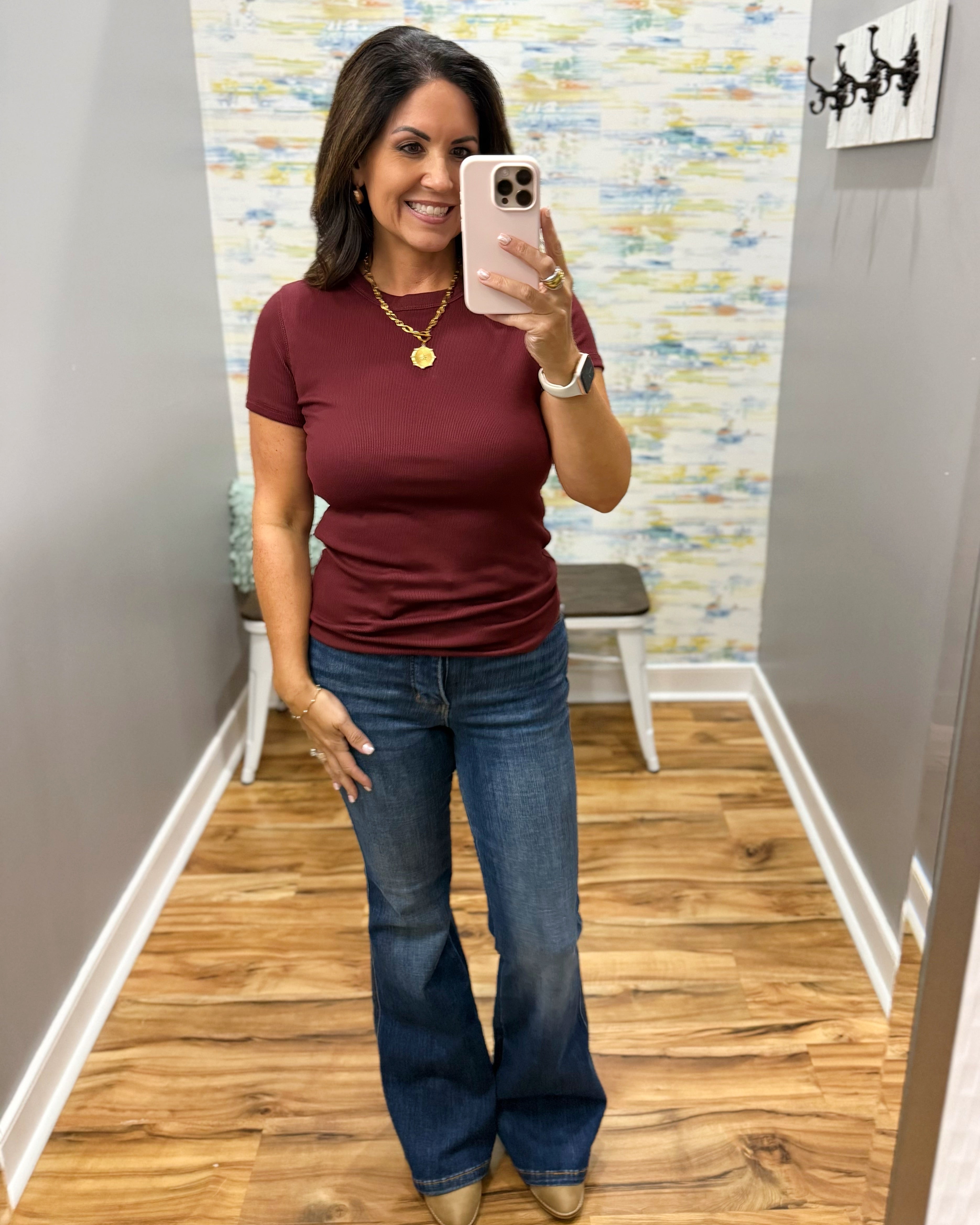 A woman takes a mirror selfie wearing a fitted short-sleeve burgundy t-shirt and blue flare jeans in a home hallway.