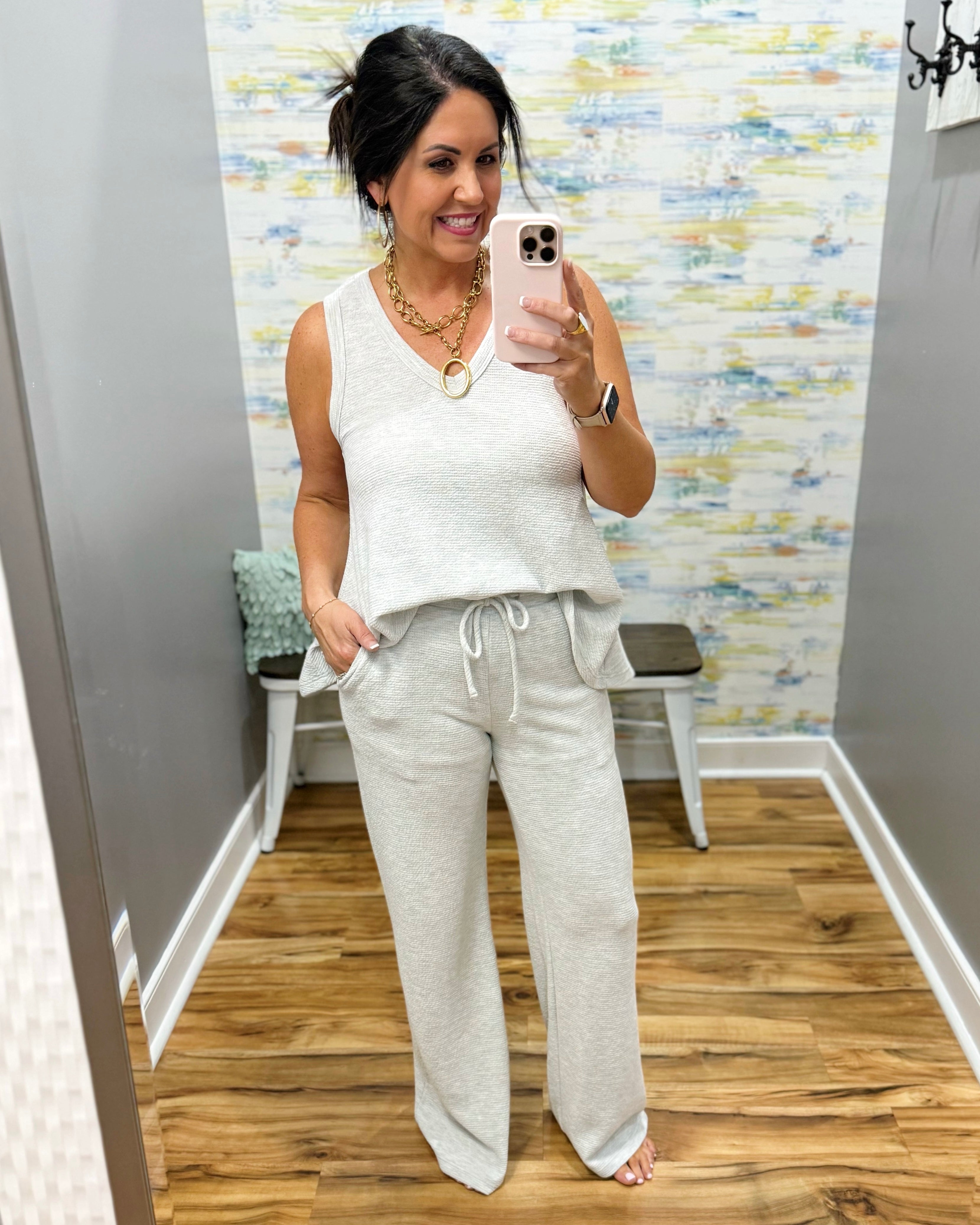 Woman taking a mirror selfie wearing a white sleeveless top and gray sweatpants in a room with a patterned wall.