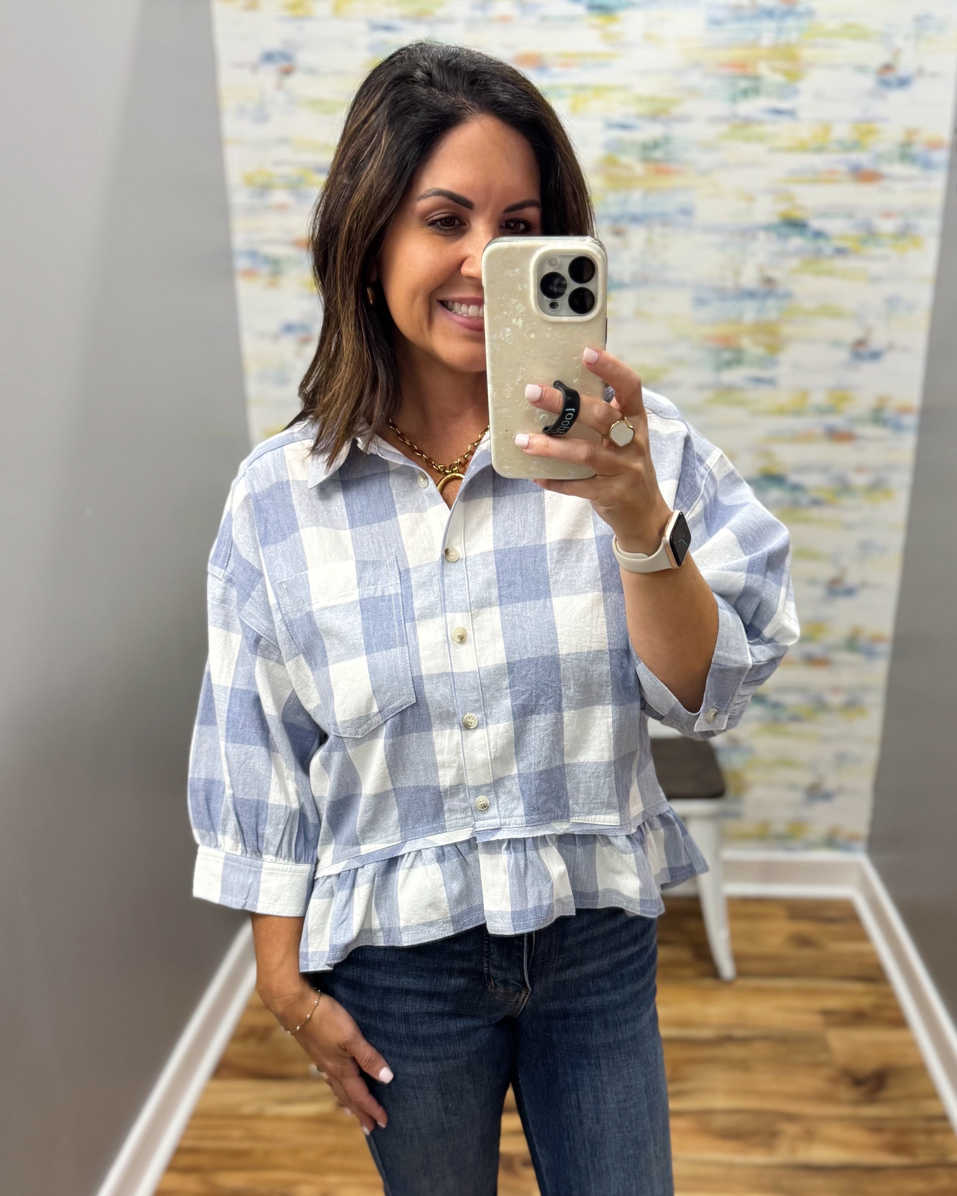 A woman taking a mirror selfie in a boutique wearing a light blue and white gingham peplum blouse with dark blue jeans.