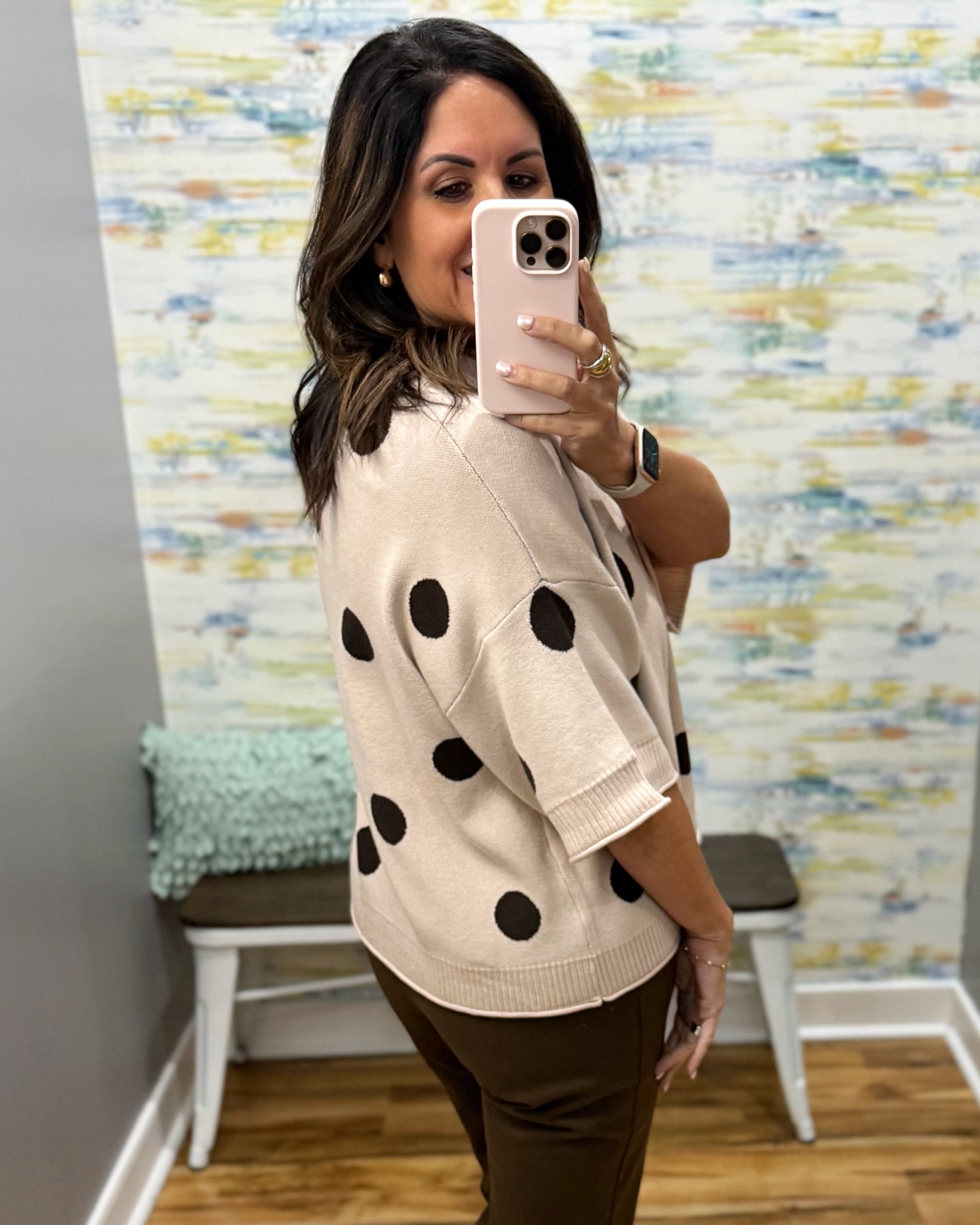 A woman takes a mirror selfie wearing a beige knit sweater with large black polka dots and brown pants in a home setting with a colorful abstract wallpaper.