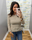 A woman taking a mirror selfie wearing a brown and cream striped long-sleeve top with blue jeans, holding a light pink smartphone.