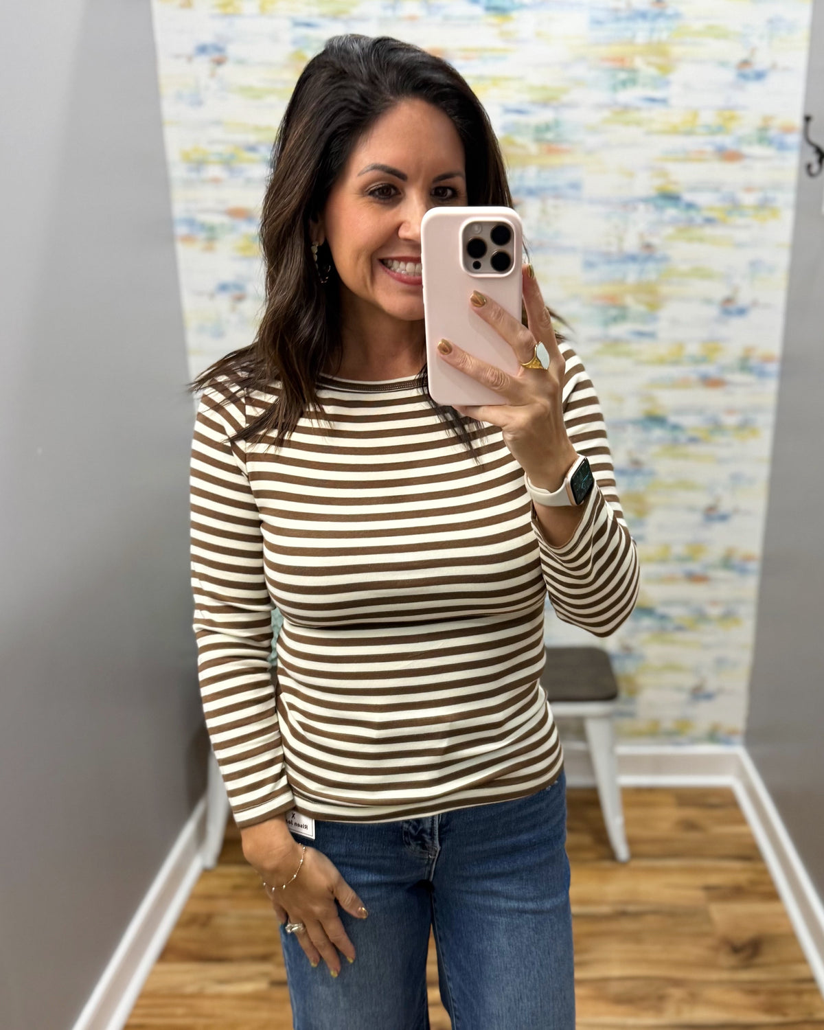 A woman taking a mirror selfie wearing a brown and cream striped long-sleeve top with blue jeans, holding a light pink smartphone.