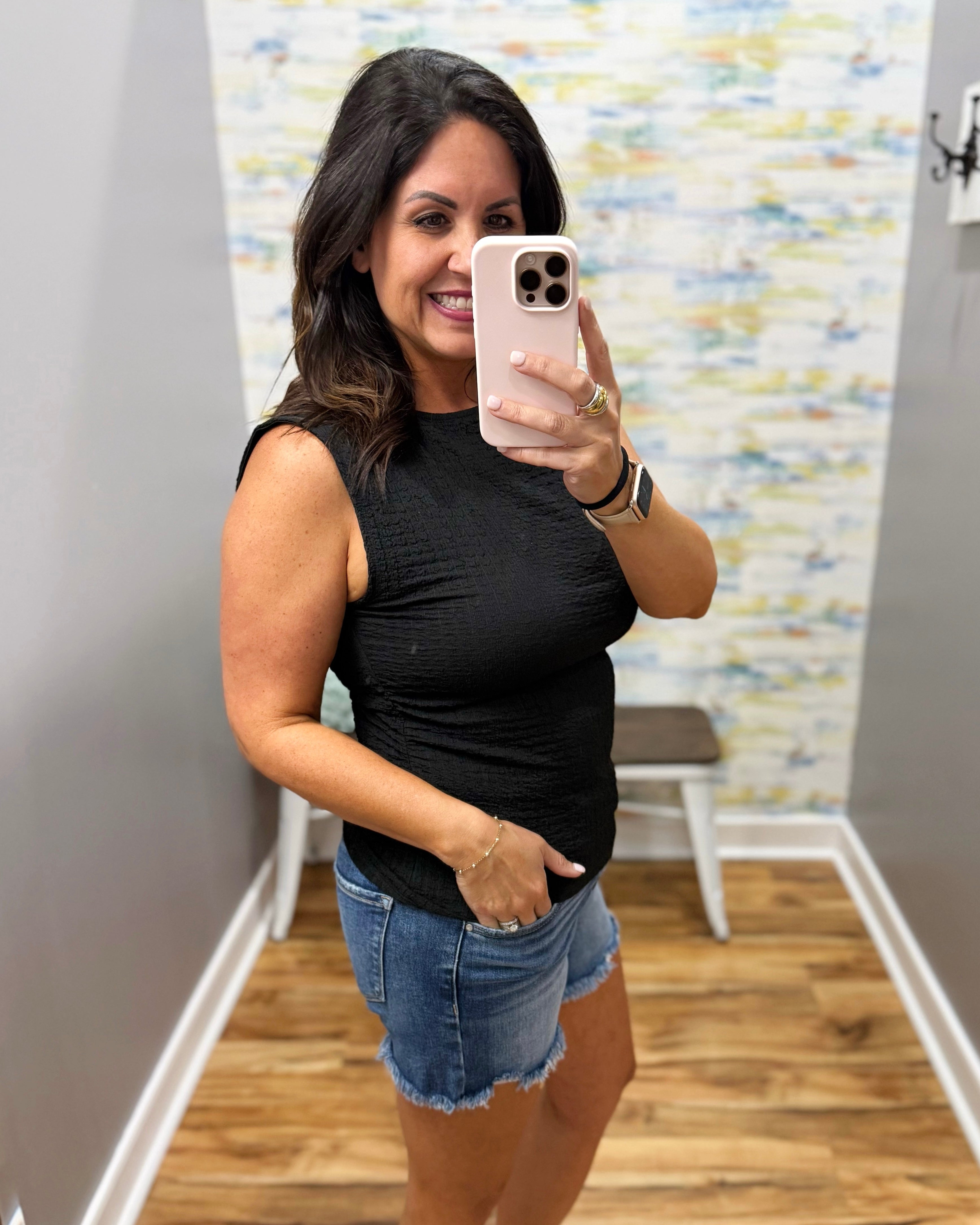 Woman taking a mirror selfie wearing a black sleeveless top and denim shorts in a room with a patterned wall.