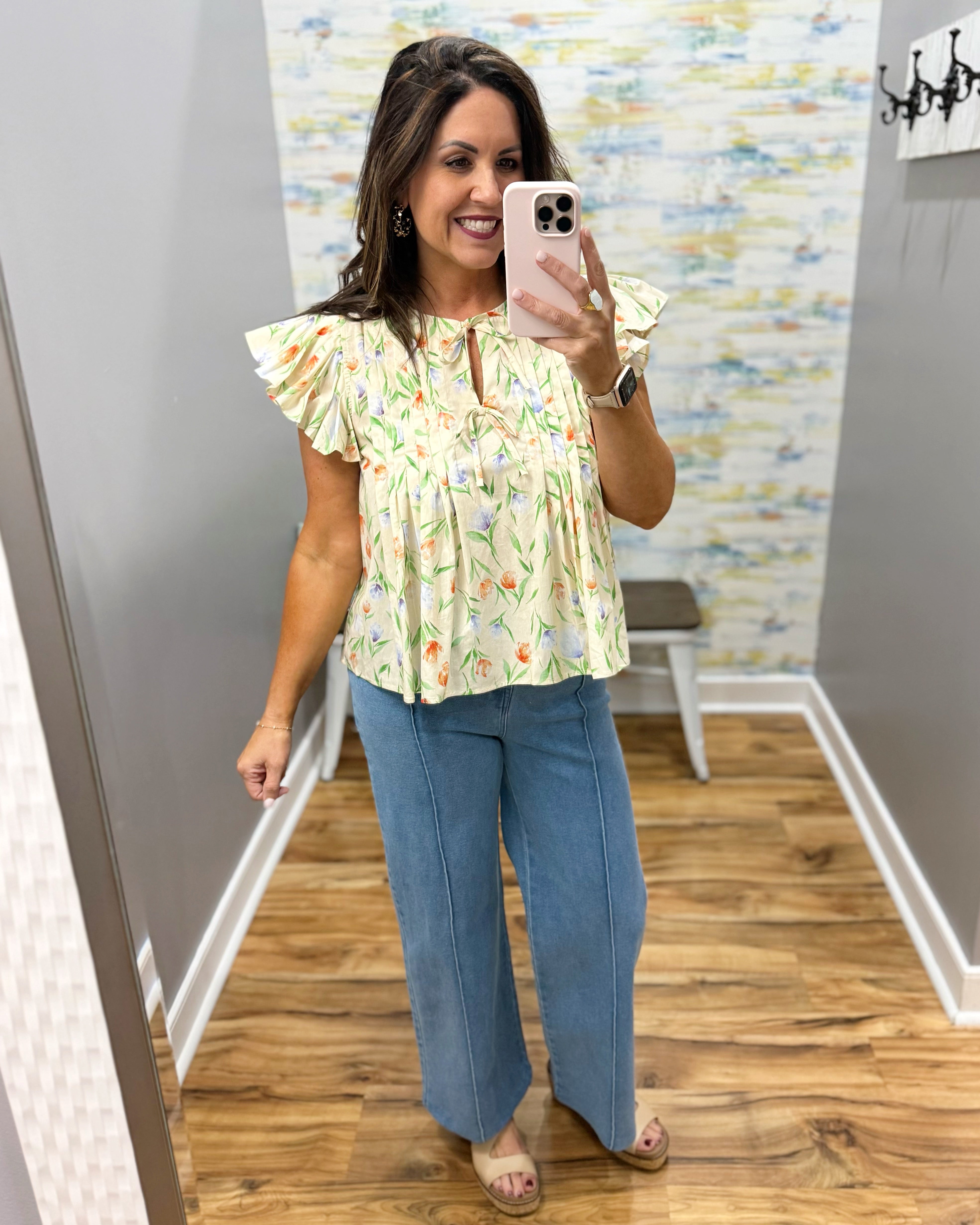 Woman taking a mirror selfie wearing a floral blouse and blue pants in a room with a colorful wall.
