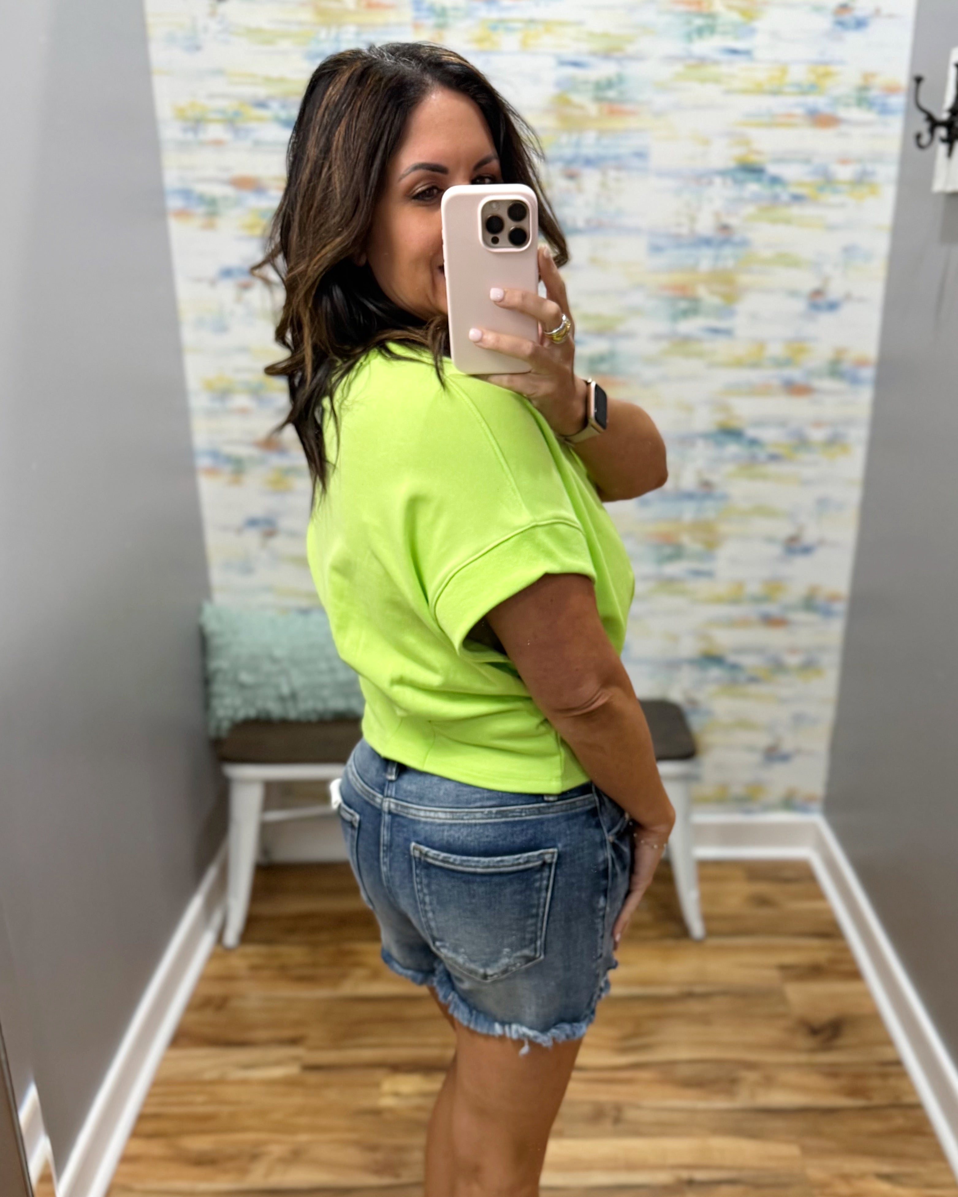 Person wearing a bright green shirt and denim shorts taking a mirror selfie.