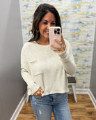 A woman takes a mirror selfie wearing a cream-colored, textured knit long-sleeve top with a front pocket and distressed blue jeans.