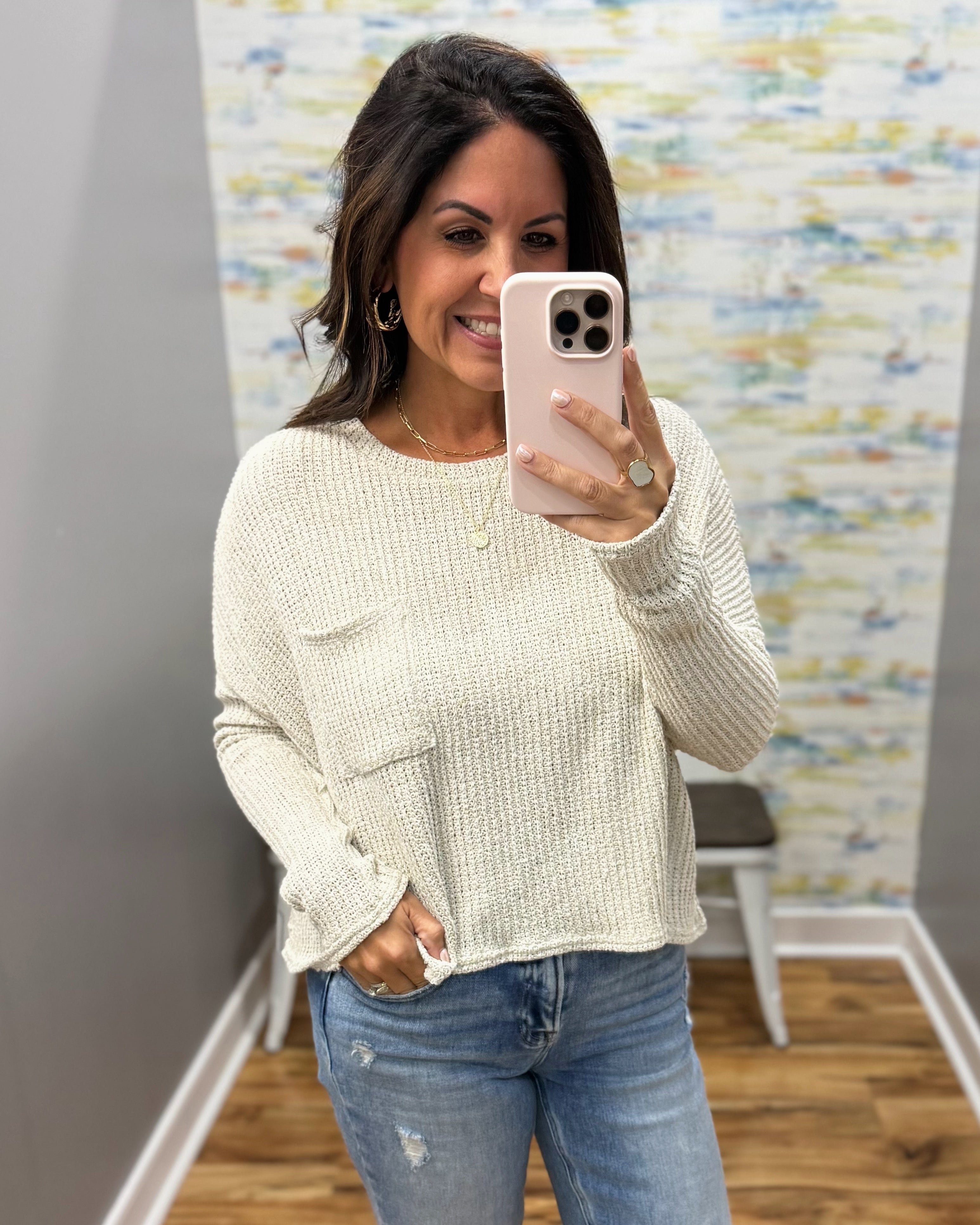A woman takes a mirror selfie wearing a cream-colored, textured knit long-sleeve top with a front pocket and distressed blue jeans.