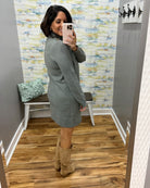 A woman takes a mirror selfie in a fitting room wearing a sage green knit cardigan dress and tan suede boots. The fitting room has gray walls, a patterned wallpaper background, a wooden floor, and a small bench with a pillow.