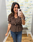 A woman taking a mirror selfie in a boutique wearing a brown short-sleeve blouse with white polka dots and blue jeans.