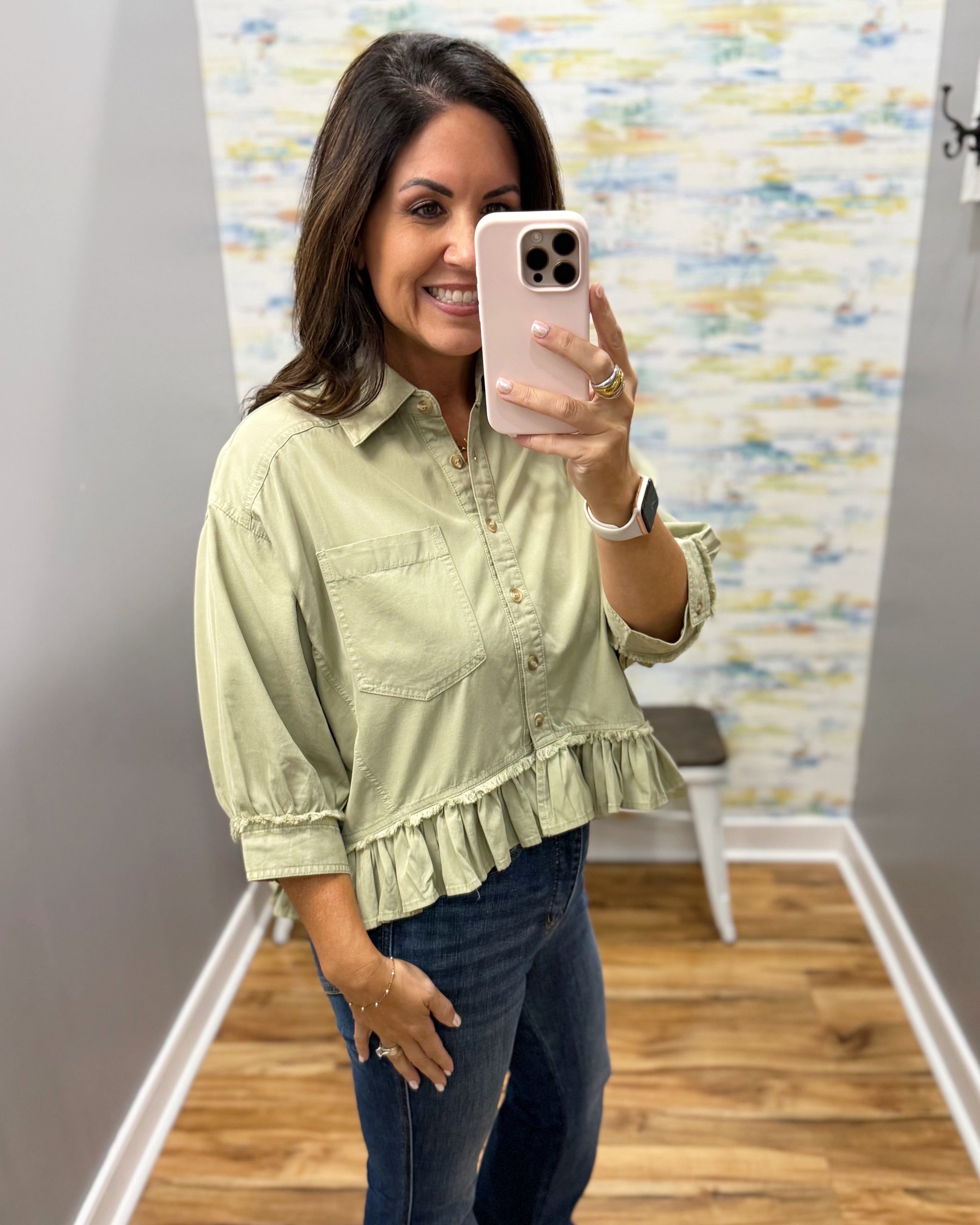 A woman takes a mirror selfie wearing a light sage green ruffled button-up blouse and dark blue jeans in a fitting room with wood floors and a patterned wall.
