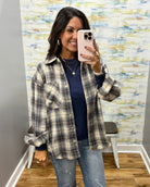 A woman takes a mirror selfie wearing an oversized plaid button-up shirt in beige, gray, and navy over a navy blue top, paired with light-wash blue jeans.