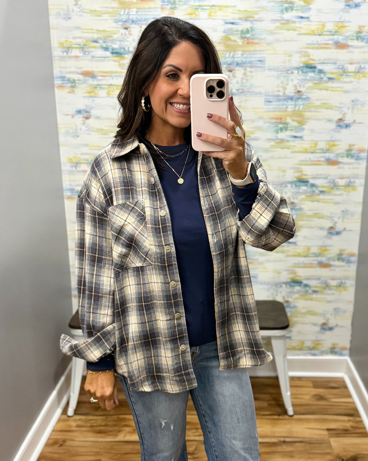 A woman takes a mirror selfie wearing an oversized plaid button-up shirt in beige, gray, and navy over a navy blue top, paired with light-wash blue jeans.
