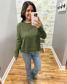 A woman takes a mirror selfie wearing an olive green long-sleeve crewneck sweater and light-wash blue jeans in a home setting with wood floors.