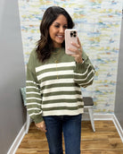 A woman takes a mirror selfie wearing an olive green and cream striped knit sweater, blue jeans, and gold necklaces in what appears to be a boutique fitting room.